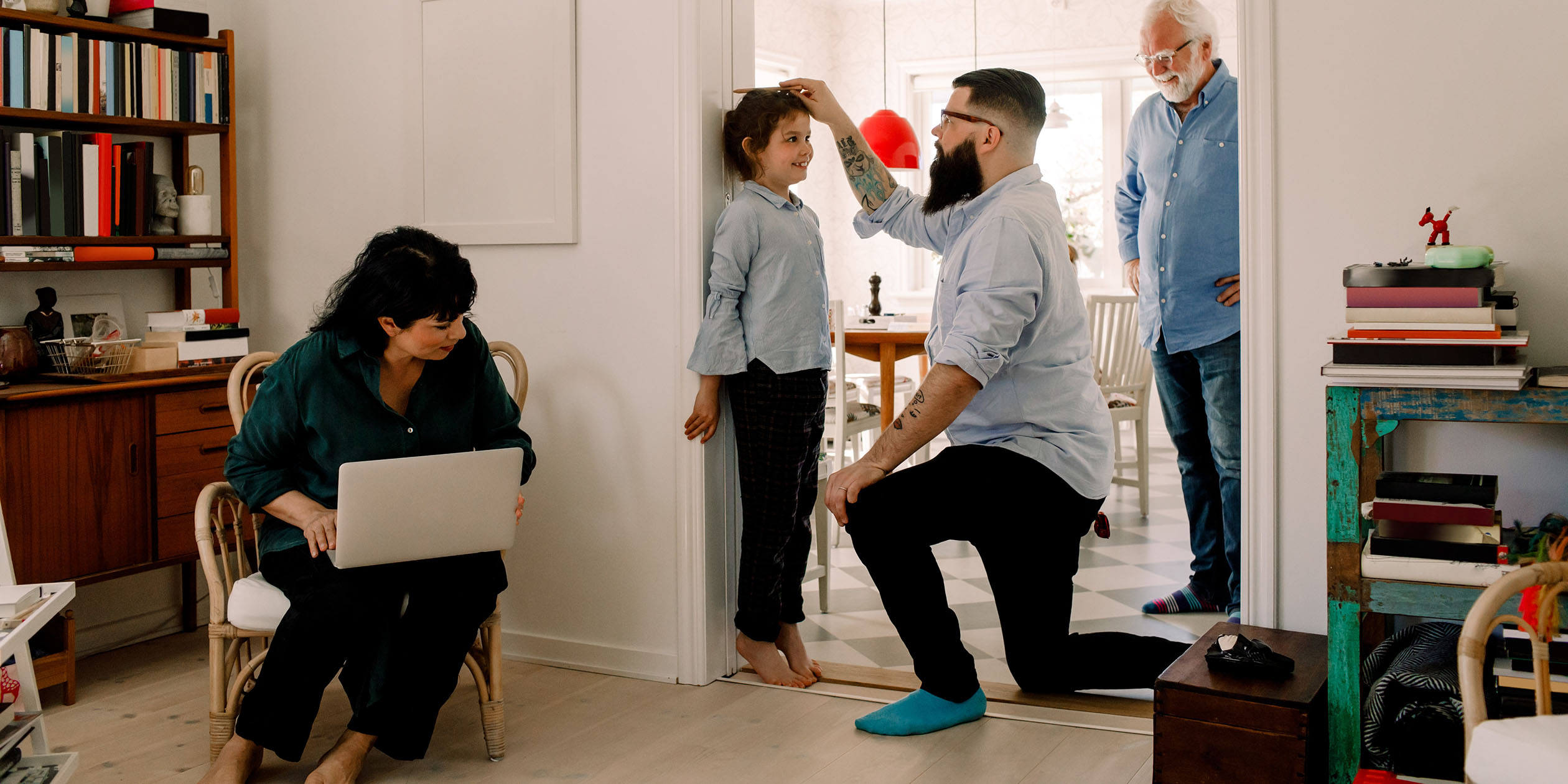 I ett utrymme mellan kök och vardagsrum står en flicka i brunt hår och ljusblå blus på tå tätt mot dörrlist för att bli mätt på hur lång hon är. Hennes pappa håller en penna i handen och markerar på dörrlisten. En äldre man i vitt hår och skägg står bredvid och tittar på. Alla på bilden ser glada ut.