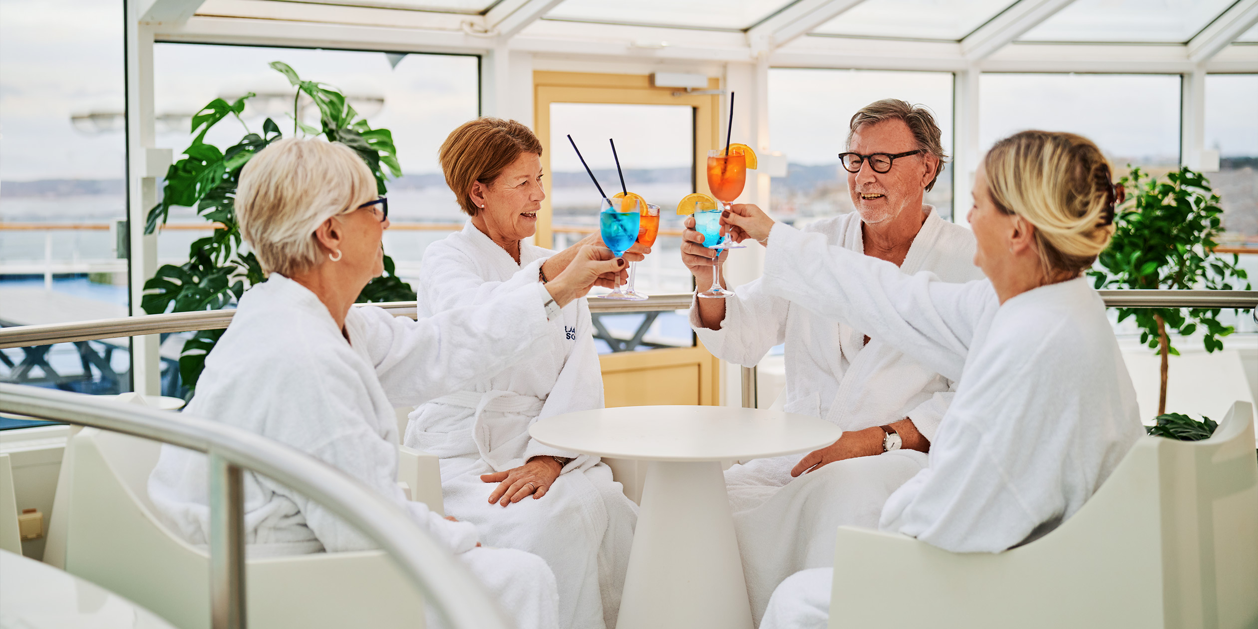 Fyra äldre män och kvinnor i vita morgonrockar skålar på ett spa.