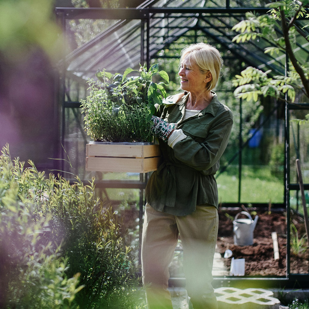 En äldre kvinna som bär på en blomlåda utanför ett växthus